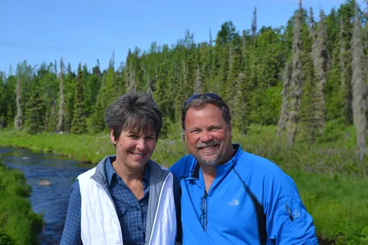 Two guests in rain gear smiling on a forested dock.